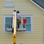 Un pintor en un ascensor pintando el revestimiento de una casa amarilla.