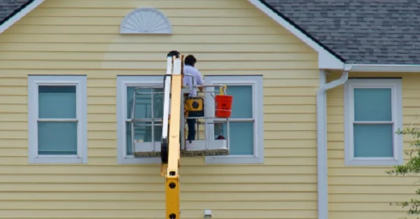 Un pintor en un ascensor pintando el revestimiento de una casa amarilla.