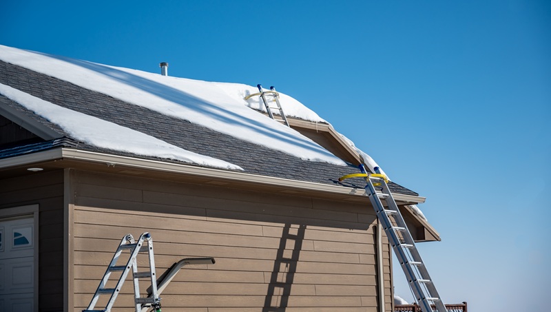 Multiple ladders setup on a residential roof covered in snow as roof is being replaced.